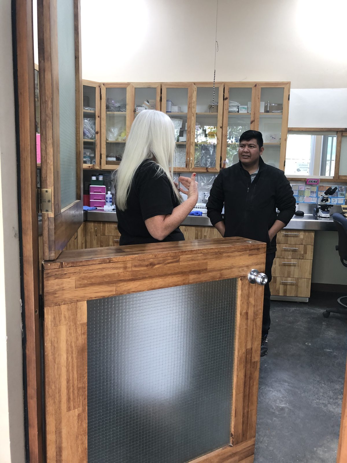 Inside the stocked Adonai medical clinic, a staff member consults with a patient beside cabinets of supplies and a microscope